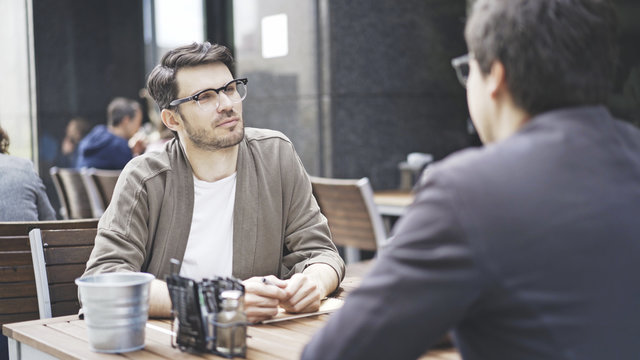 Two friends are talking having a conversation at table of the cafe outdoors. A man dressed in a jacket wearing eyeglasses listening to his friend talking