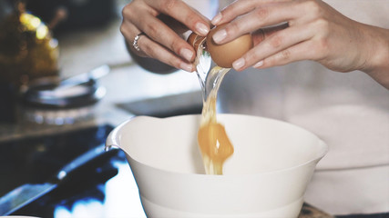 A young woman dressed in a white shirt is breaking an egg into a white bowl. A young woman is cooking in a modern kitchen.