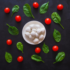 Italian mozzarella cheese, cherry tomatoes and spinach leaves on a black table. Pattern.