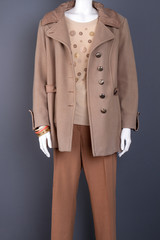 Female fashionable autumn overcoat. Mannequin dressed in female beige blouse and brown trousers, grey background. Boutique of women clothes.
