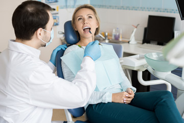 Doctor in gloves and mask exploring teeth of young girl with small mirror while sitting in chair. 