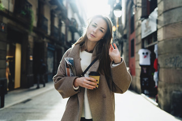 Charming young journalist going to the work with take away coffee and newspaper in her hand on a sunny morning. Office worker female wearing casual clothes enjoying coffee after working day