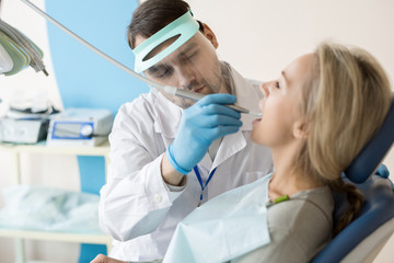 Confident man in protective mask drilling tooth with machine working with young woman in chair. 