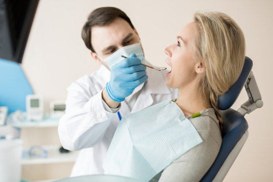 Side view of girl sitting in chair while dentist with small mirror exploring teeth conditions. 