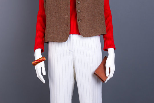 Mannequin with fashion clothes and accessories. Female mannequin dressed in red sweater, brown waistcoat and white trousers. Dummy with bracelet and wallet.