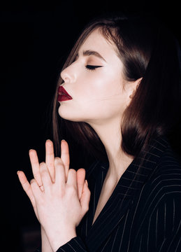 Pray and hope concept. Girl on calm face with closed eyes in black jacket, black background. Woman with stylish makeup and hairstyle holds hands as praying, side view.
