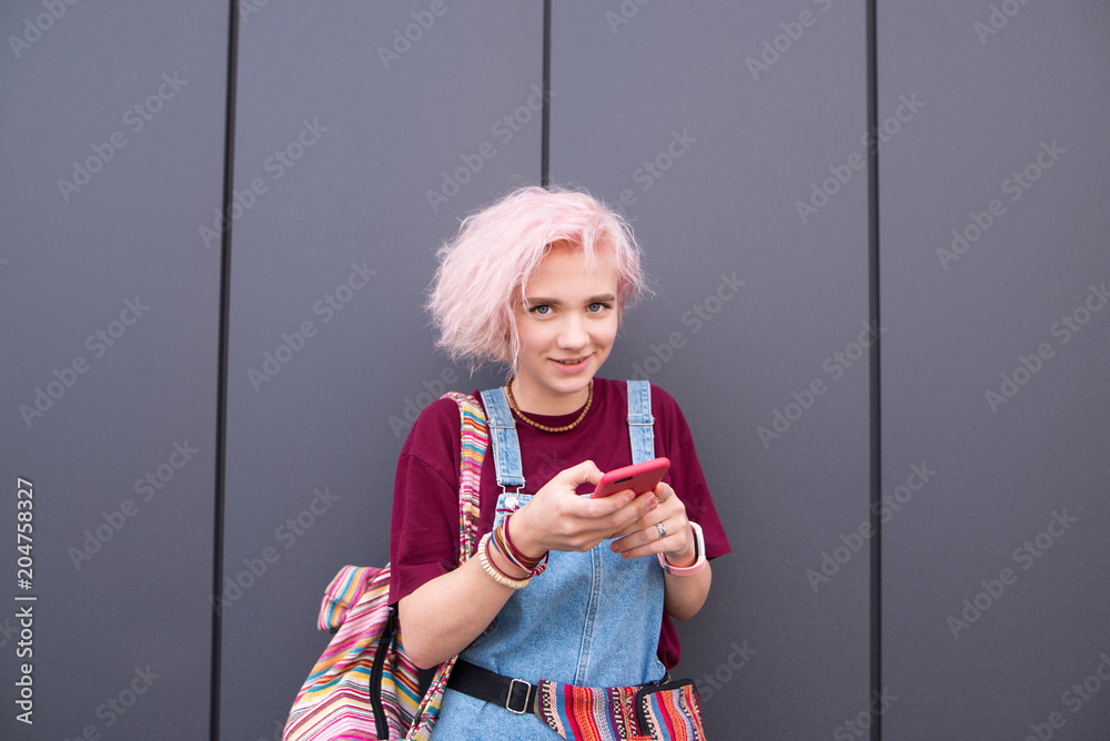 Wall mural Stylish happy girl with colored hair is with a smartphone on a dark background. Smiling girl hipster with a smartphone in her hands looks at the camera on the background of a black wall