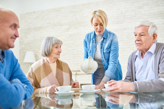 Woman Pouring Tea For Senior Patients In Assisted Living Home Sitting At Table And Chatting. 