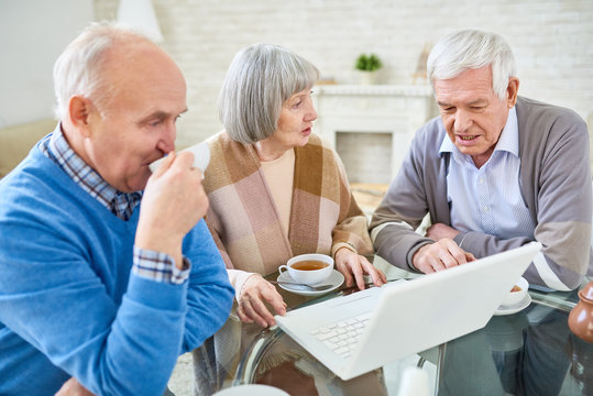 Elegant senior woman and men drinking tea and surfing laptop together sitting at table in assisted living home. 