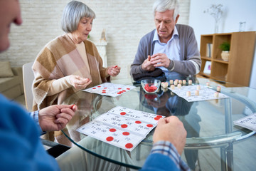 Senior patients of assisted living home gathering at table in living room and playing lotto at table. 