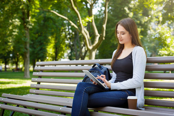 Obraz premium Motivated asian freelancer in the park. Girl on a bench, drinking coffee and working with tablet