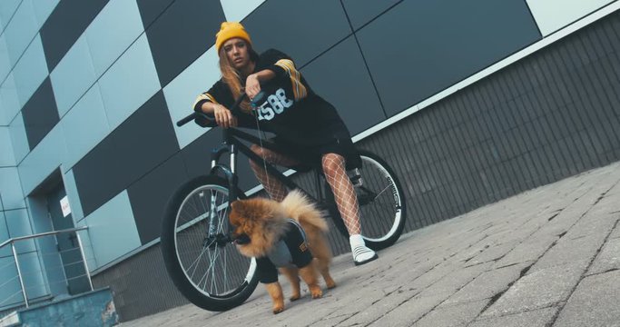 Young stylish Caucasian female posing on a bicycle with her Pomeranian Spitz dog puppy outdoors. 4K UHD RAW edited footage