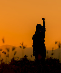 Silhouette woman at sunset standing elated with arms raised up above her head.