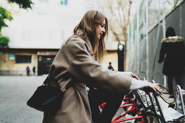 Stylish student girl in stylish clothes sitting on a rented city bicycle and ready to ride on. Freelancer female renting the bike to explore the city. Young tourist girl choosing bike to ride around