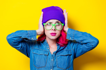 Young pink hair girl in blue shirt, purple hat and green glasses. Portrait isolated on yellow background