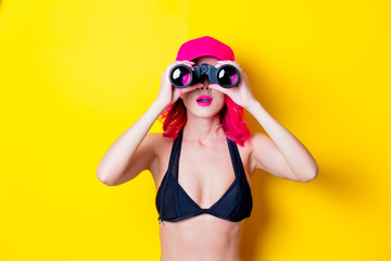 Young pink hair girl in bikini with cap and binoculars. Portrait isolated on yellow background