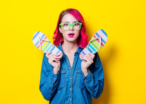 Young pink hair girl in blue shirt and green glasses holding a colored flip flops sandals. Portrait isolated on yellow background