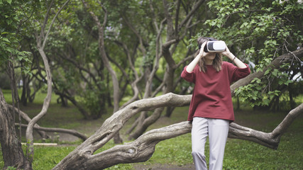 A young girl dressed in a red sweatshirt and gray pants having fun in in virtual reality glasses sitting on a tree trunk in the park