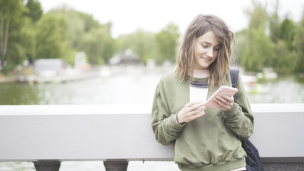 Fototapeta premium A smiling happy laughing young girl is listening to a music outdoors dressed in a green sweatshirt drinking a cup of coffee