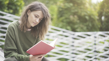 A closeup of a concentrated young casual brunette girl dressed in a green sweat shirt is sitting on the stairs holding a book on the stairs