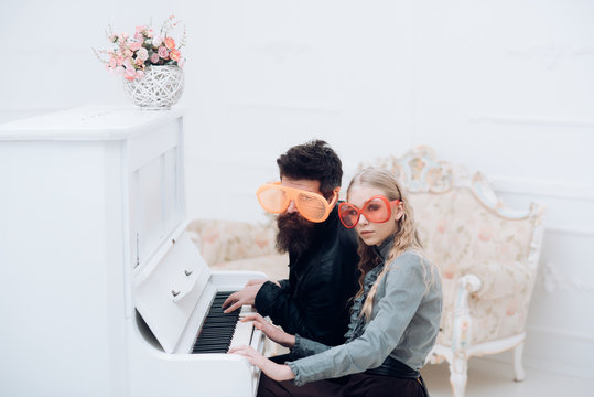 Young couple in enormous glasses sitting in white room and playing piano. Stylish beaded man and his blond girlfriend sitting at grand piano