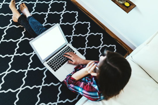 Student girl sitting on a carpet with a laptop on her knees with blank white display for your content. Freelancer female calling to clients by mobile phone. Hipster girl talking by smartphone at home