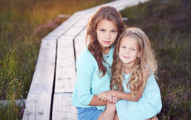 Fototapeta premium Sisters relaxing outdoors while sitting on a wooden path in a field at sunset.