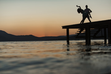 Silhouette of sensual couple dancing on pier with sunset above sea surface on background. Romance and love concept. Couple in love on romantic date in evening at dock, copy space.