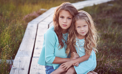 Fototapeta premium Sisters relaxing outdoors while sitting on a wooden path in a field at sunset.