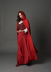 Naklejka premium full length portrait of woman wearing red fantasy costume with cloak, standing pose on grey studio background.