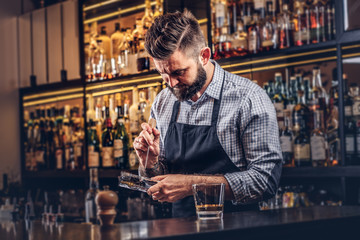 Stylish bearded barman in a shirt and apron splits ice to make a cocktail at bar counter background.