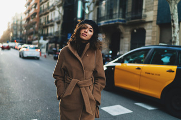 Fototapeta premium beautiful girl with long curly hair in a brown coat takes a taxi standing on the transition passage of a street in a European city. modern businesswoman walks around Barcelona during a business trip