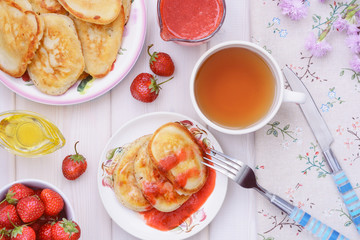 Fresh pancakes, strawberries, strawberry sauce, honey, a cup of tea are on a wooden table ready for tea drinking