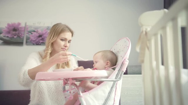 Beautiful mom try feeding baby with porridge. Blondy woman feed kid in baby chair at home. Family care. Infant gnawing teethers