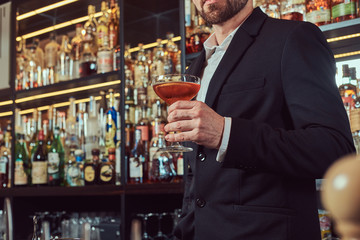 Handsome stylish male holds a glass of exclusive alcohol standing at bar counter background.