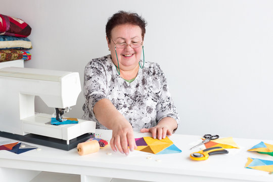 work, manufacturing, quilting concept. there is an old and professional seamstress at her work table with all tools that she needs and sewing machine, she is holding bright patches and smiling