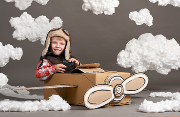 the boy plays in an airplane made of cardboard box and dreams of becoming a pilot, clouds of cotton wool on a gray background
