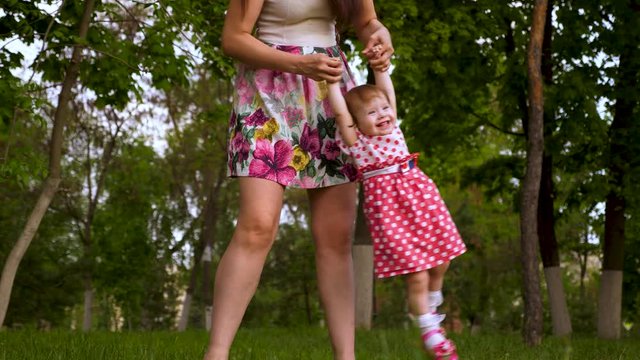 Mom is shaking hands in air with a happy laughing daughter. Parent plays with baby on green grass in summer park. Happiness of motherhood. Rest in open air.