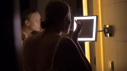 Young woman looking in face cosmetic mirror in the bathroom after applying night cream