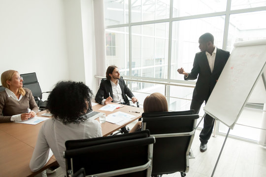 African business coach gives presentation at board meeting working with flipchart, executive team sit at conference table listening to black speaker presenting new marketing sales project in office