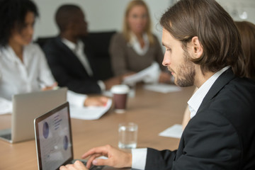 Fototapeta premium Serious businessman in suit working on laptop online at diverse group boardroom meeting, executive project manager using computer business software for statistic data analysis at multiracial briefing