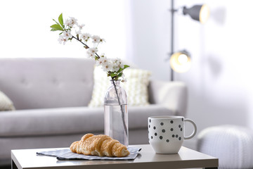 Fresh croissant, cup and flowers on table in room