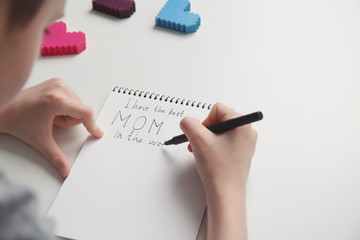 Little child writing congratulation for Mother's Day in notebook on light background