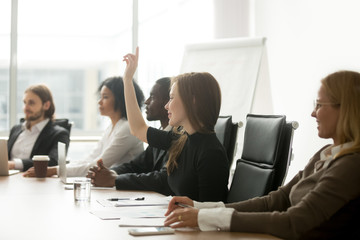 Smiling curious young businesswoman raising hand at multiracial group meeting engaging in offered...