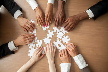 Hands of diverse people connecting puzzle together on office desk, multi-ethnic team engaging in...