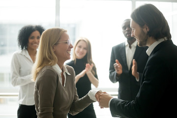 Boss handshaking rewarding motivating successful female employee congratulating with promotion, appreciating good work results, making partnership deal, expressing respect while colleagues applauding