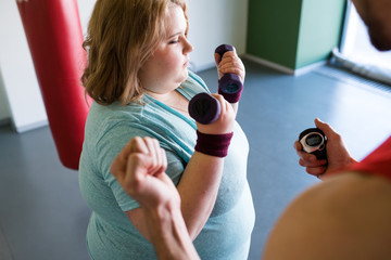 High angle portrait of obese woman exercising with dumbbells during weightloss training in gym with...