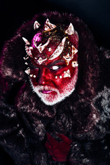 Senior man with white beard dressed like monster. Alien, demon, sorcerer makeup. Evil concept. Man with thorns or warts in fur coat. Demon with red face on black background, close up.