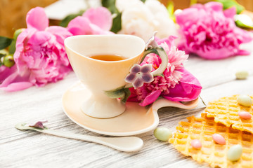 Fototapeta na wymiar Cup of green tea and spring peonies blossom on a old wooden background with waffles and caramels. Rustic.