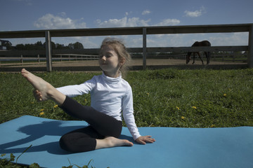 Fototapeta na wymiar The young girl is engaged in gymnastics on the grass healthy lifestyle. Child girl doing gymnastics. Little cute girl practicing yoga pose on the background of the paddock with a horse